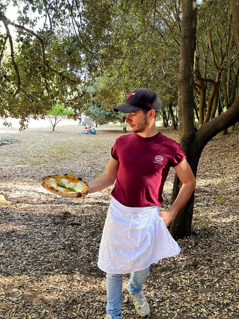 Alessio Oliva all'aperto, con grembiule bianco e una pizza appena sfornata in mano