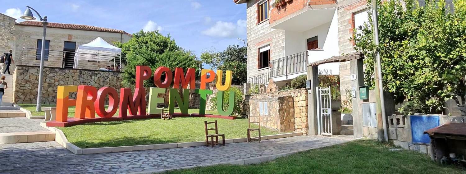 La grande scritta colorata POMPU FROMENTU sul prato della piazza di Pompu